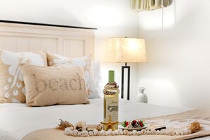 Cama arrumada com almofadas decorativas, uma garrafa de vinho e um prato de petiscos em uma mesa de cabeceira.