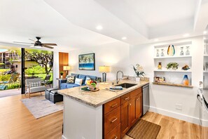A modern kitchen with a central island, wooden cabinets, and a view of a lush garden. - Papakea Villas | Ocean View 1 BR | Car Incl. w/ 6+ Nights | PKD-104 by KBM (Lahaina)