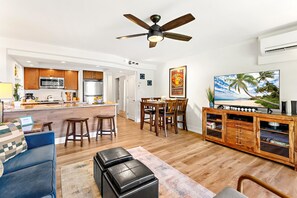 Uma cozinha moderna com ilha central, sala de jantar com cadeiras de madeira, sala de estar com sofá azul e TV sobre apoio de madeira.