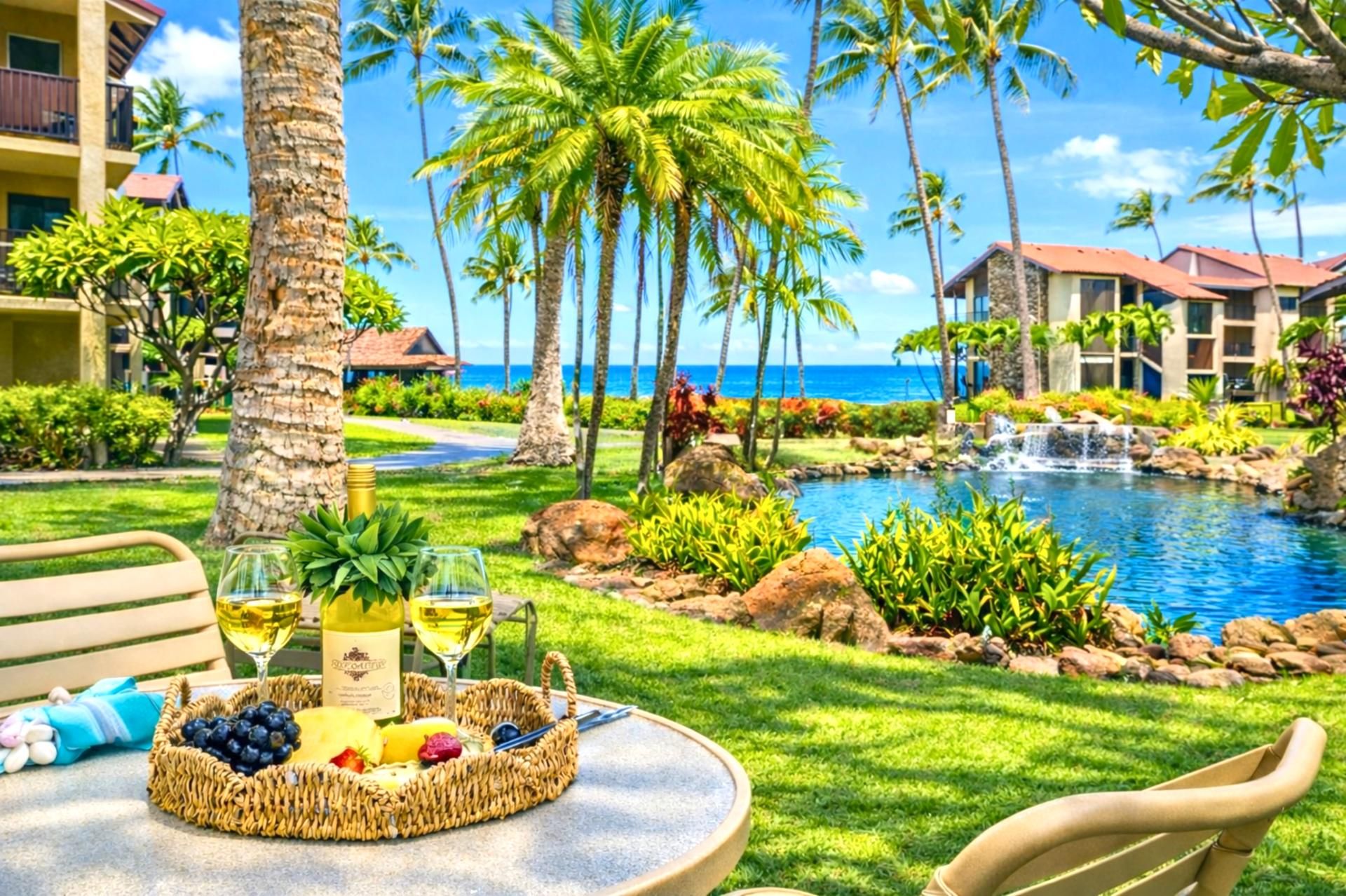 Un resort tropical con piscina, palmeras y un comedor con una cesta de frutas y copas de vino.