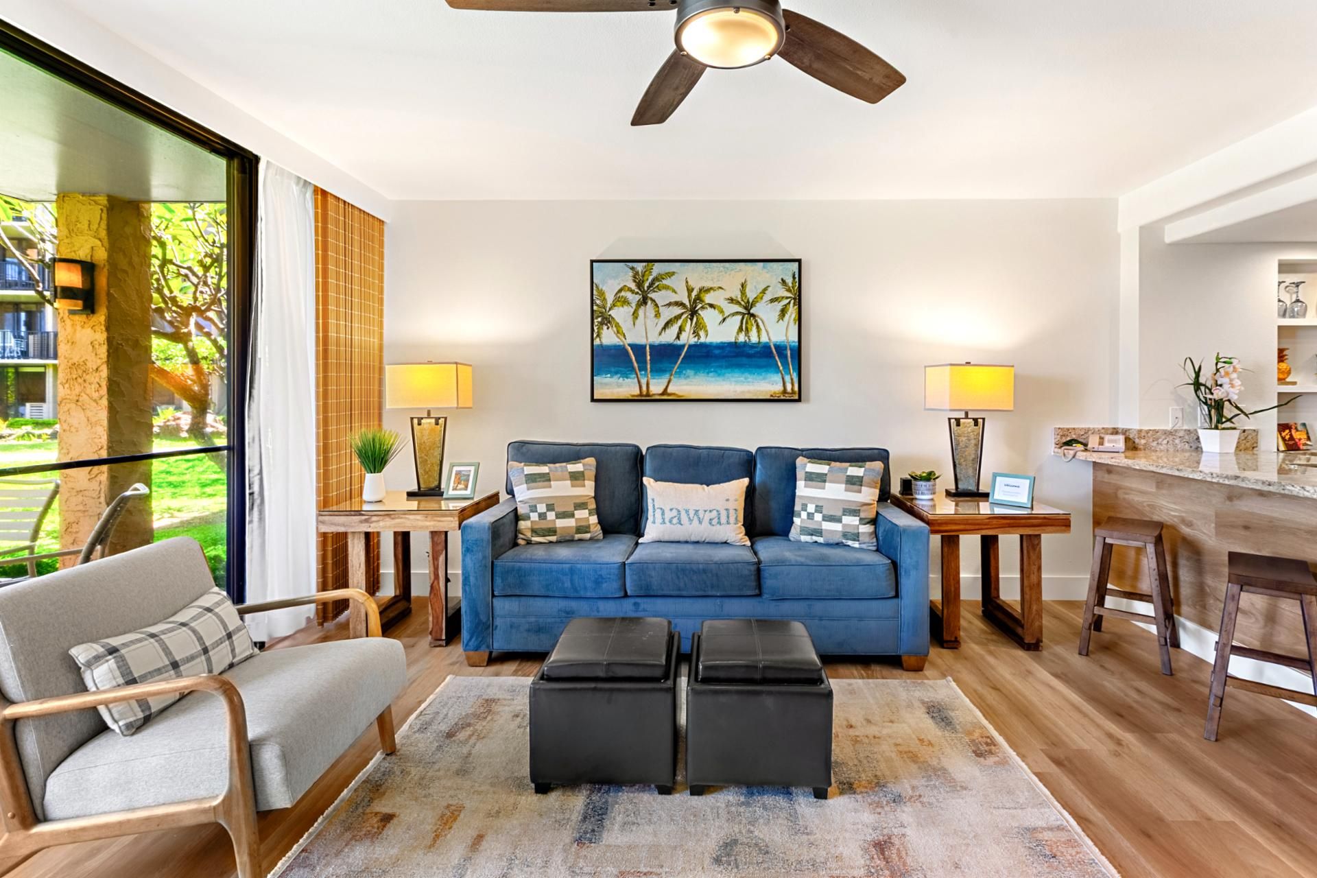 Uma sala de estar com um sofá azul, dois pufes pretos, uma mesa de centro de madeira, uma pintura de palmeiras e um ventilador de teto.