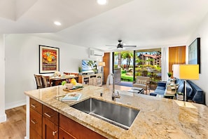 A modern kitchen with a large island, a dining area with a table and chairs, a living area with a sofa and a TV, and a view of a tropical landscape through large windows.