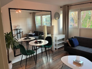 A modern living room with a sofa, a dining table with chairs, a small white desk, and a large mirror.