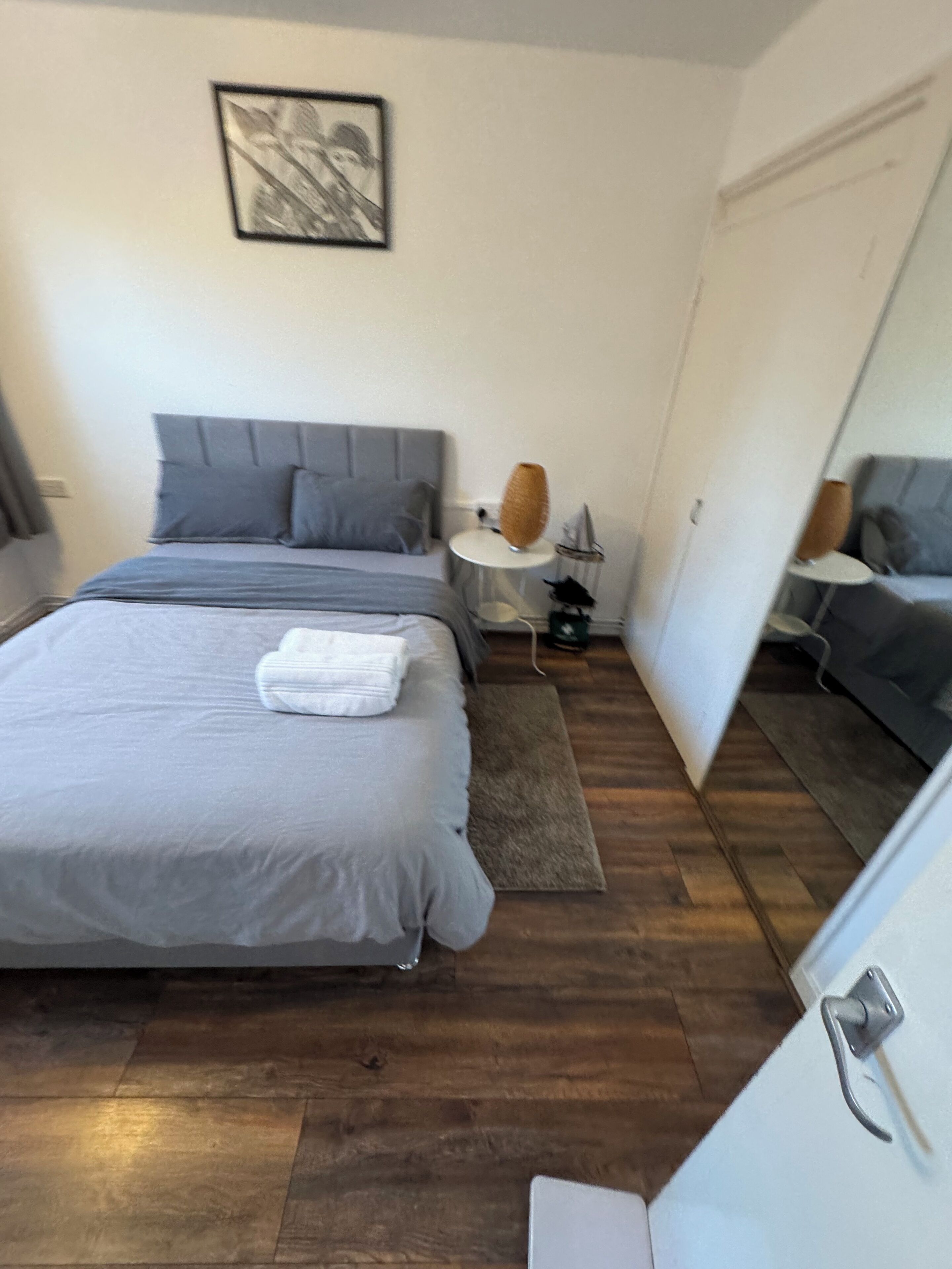A bedroom with a bed, a bedside table, a lamp, a mirror, and a door.
