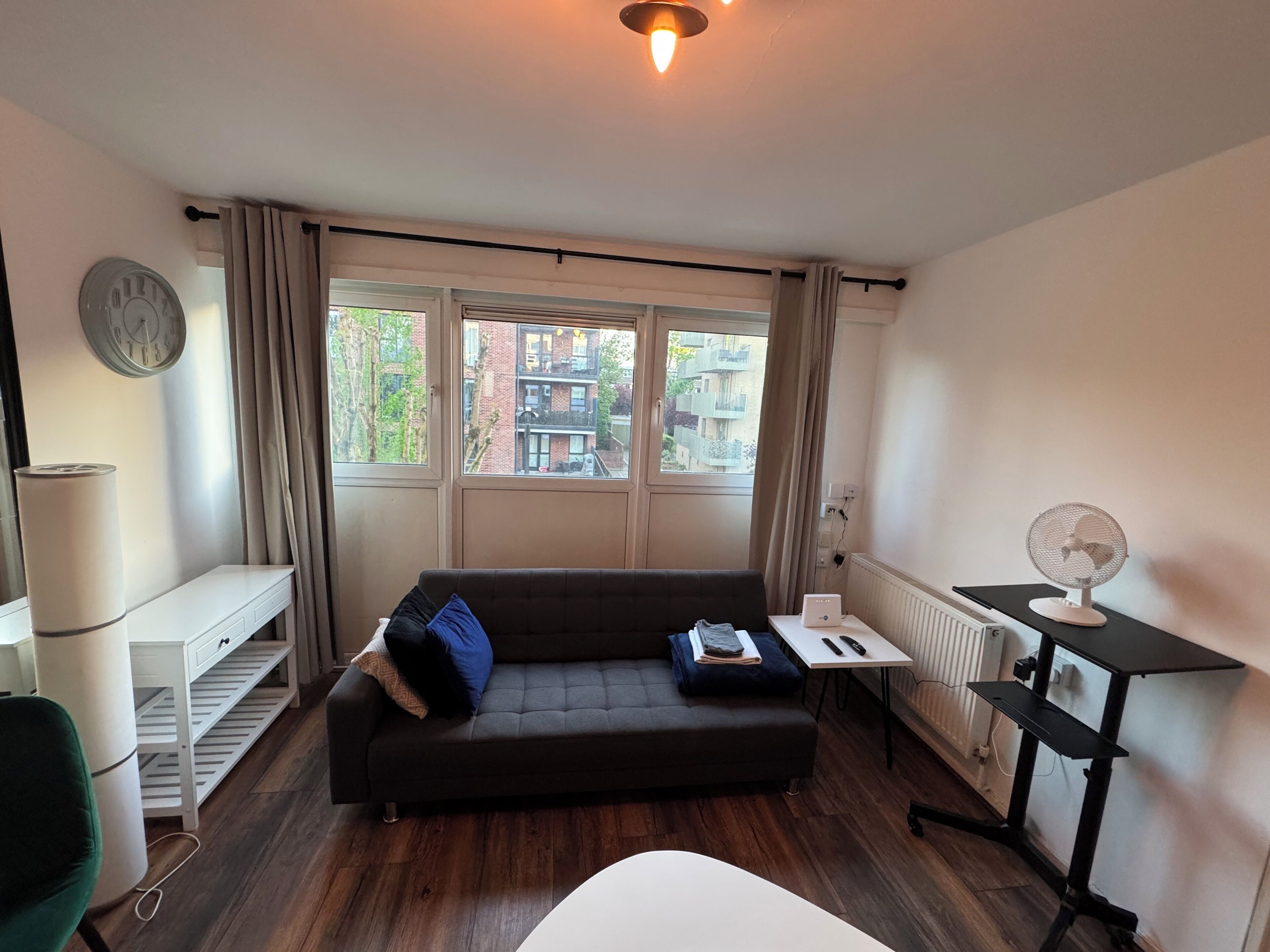 Ein Zimmer mit einer Couch, einem kleinen Tisch mit Ventilator, einem Heizkörper und einem Fenster mit Vorhängen.
