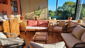 Uma sala de estar aconchegante com móveis de vime, uma estante de livros e vista para o verde lá fora.
