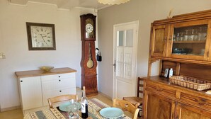 Uma sala de jantar com uma mesa posta para dois, um armário de madeira, um relógio de parede e uma porta com painéis de vidro jateado.