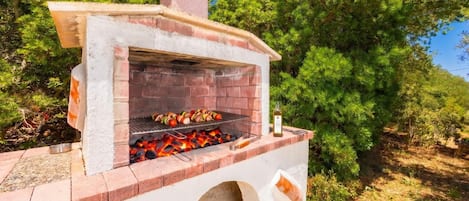 An outdoor brick oven grill with food being cooked, surrounded by trees.