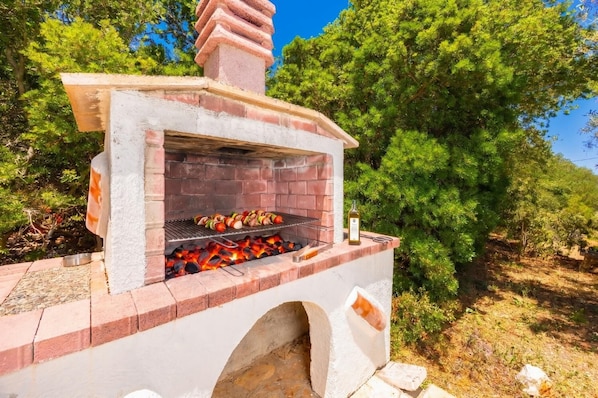 Una parrilla de ladrillo al aire libre con comida cocinándose, rodeada de árboles.