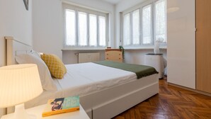 A bedroom with a bed, bedside table, lamp, and a book. There are windows with curtains and a radiator. - Casa Gabel by Wonderful Italy (Torino)