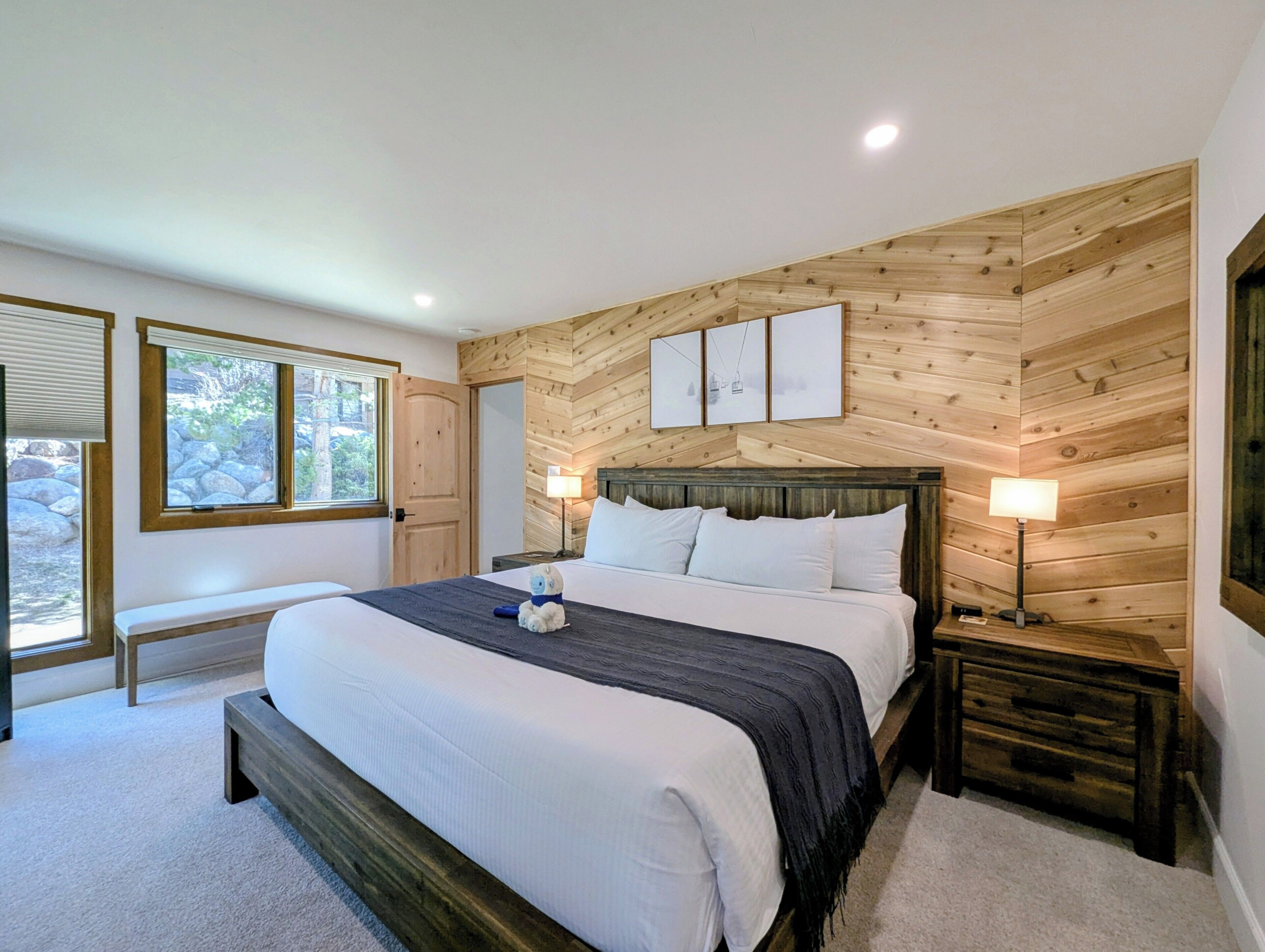 A bedroom with a large bed, wooden panel walls, a bench, a bedside table, and a window with a view of rocks and greenery.