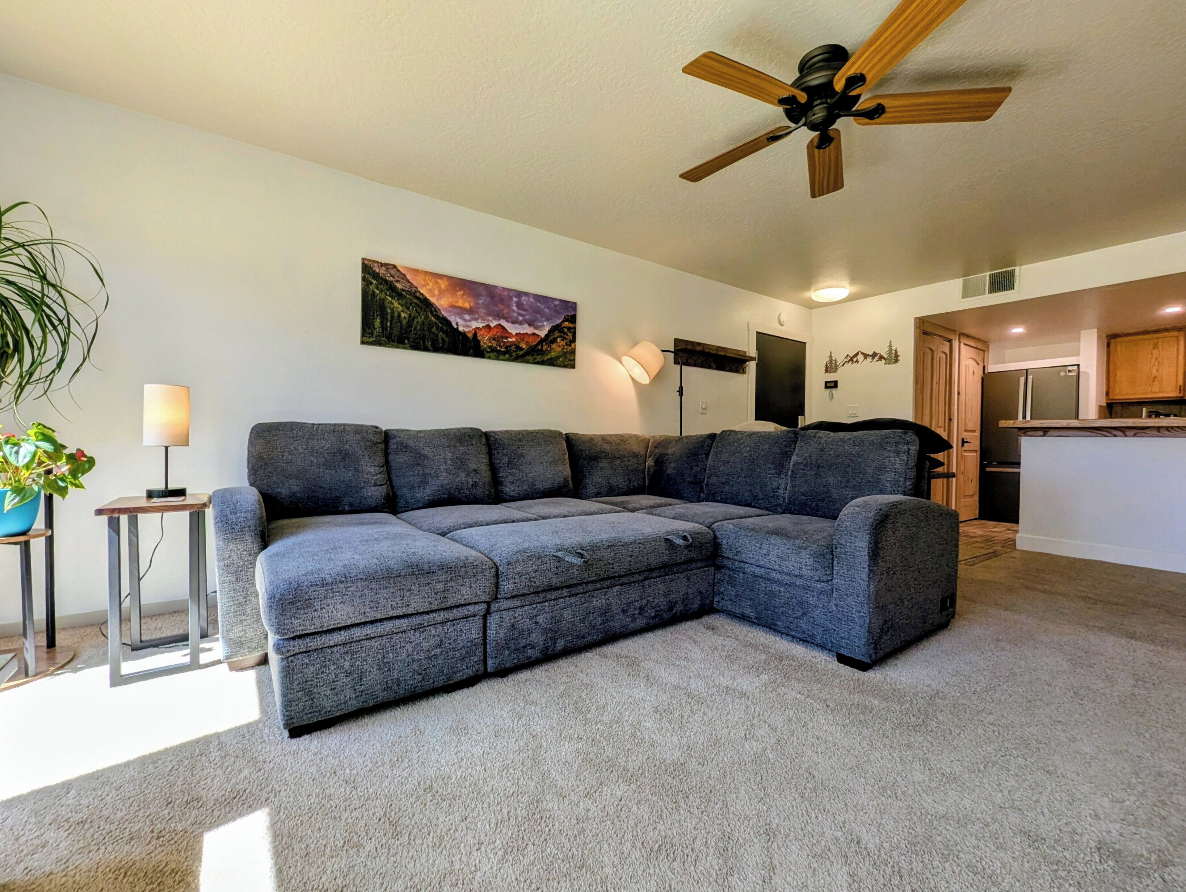 A living room with a sectional sofa, a ceiling fan, a painting, a lamp, and a kitchen area.