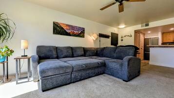 A living room with a sectional sofa, a ceiling fan, a painting, a lamp, and a kitchen area.