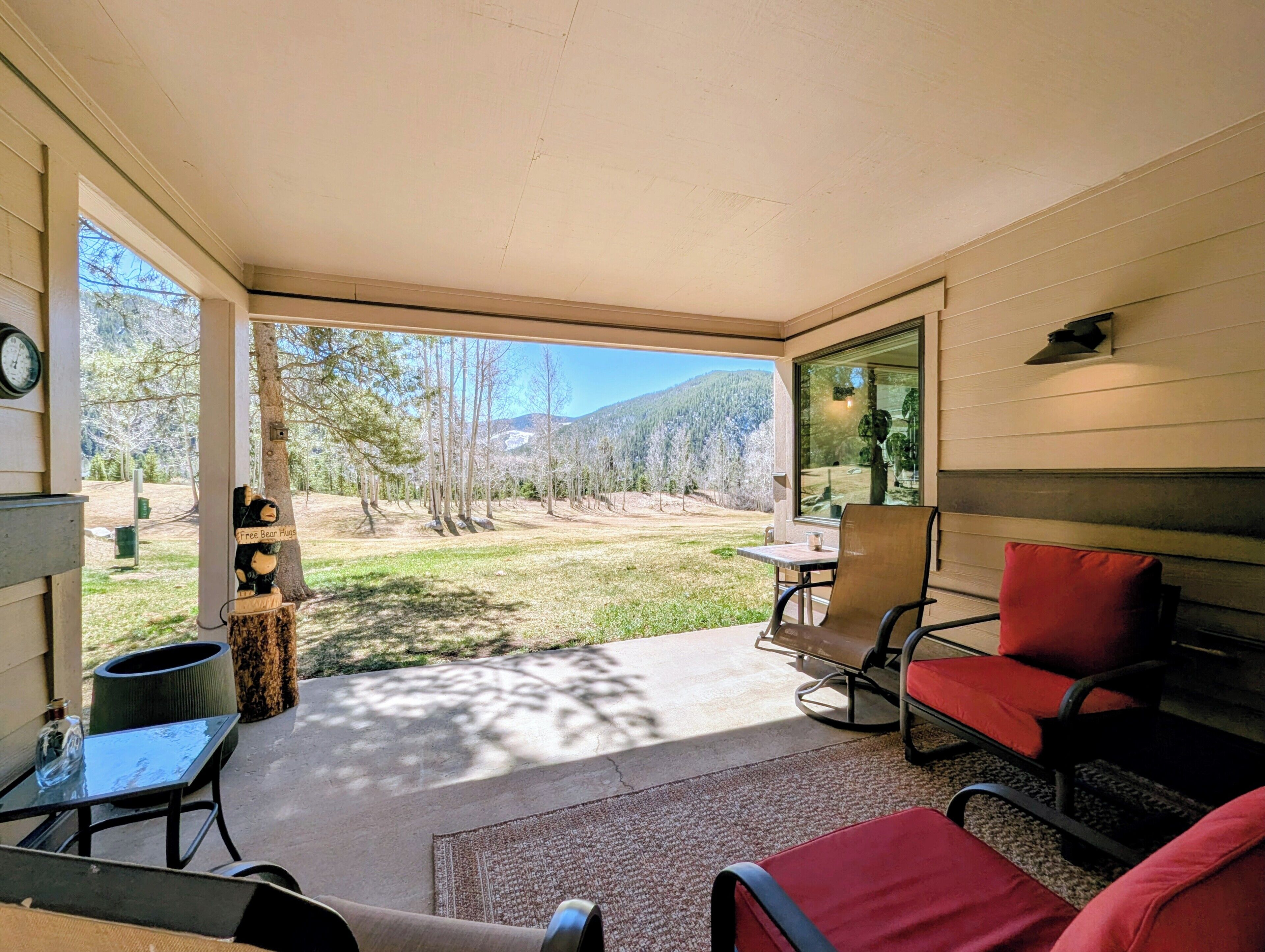 A covered patio with a view of a grassy area and trees, a table with chairs, and a clock on the wall.