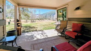 Une terrasse couverte donnant sur une pelouse et des arbres, une table avec des chaises et une horloge au mur.