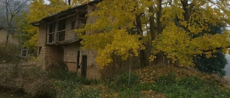 A rustic stone building with a wooden balcony, surrounded by autumn foliage and a small body of water.
