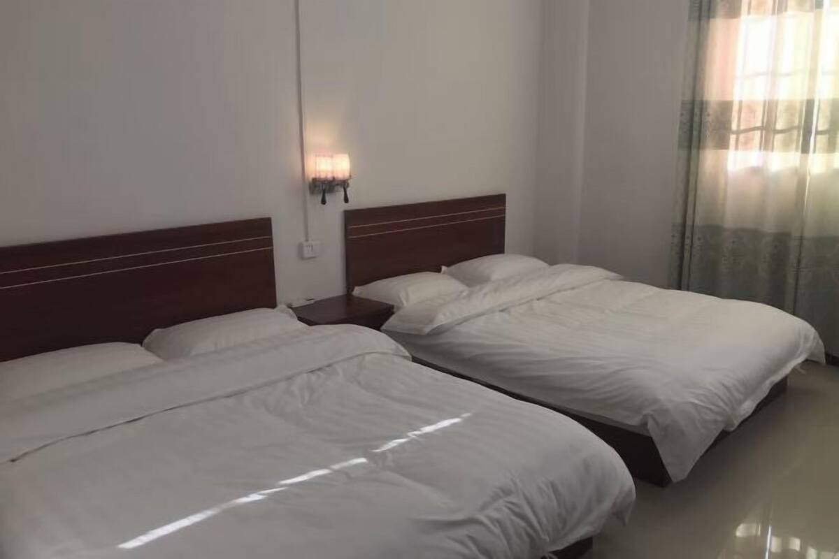 Une chambre d’hôtel avec deux lits simples, une tête de lit en bois, une table de chevet avec une lampe et une fenêtre avec des rideaux légers.