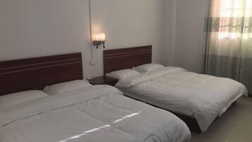 A hotel room with two single beds, a wooden headboard, a nightstand with a lamp, and a window with sheer curtains.