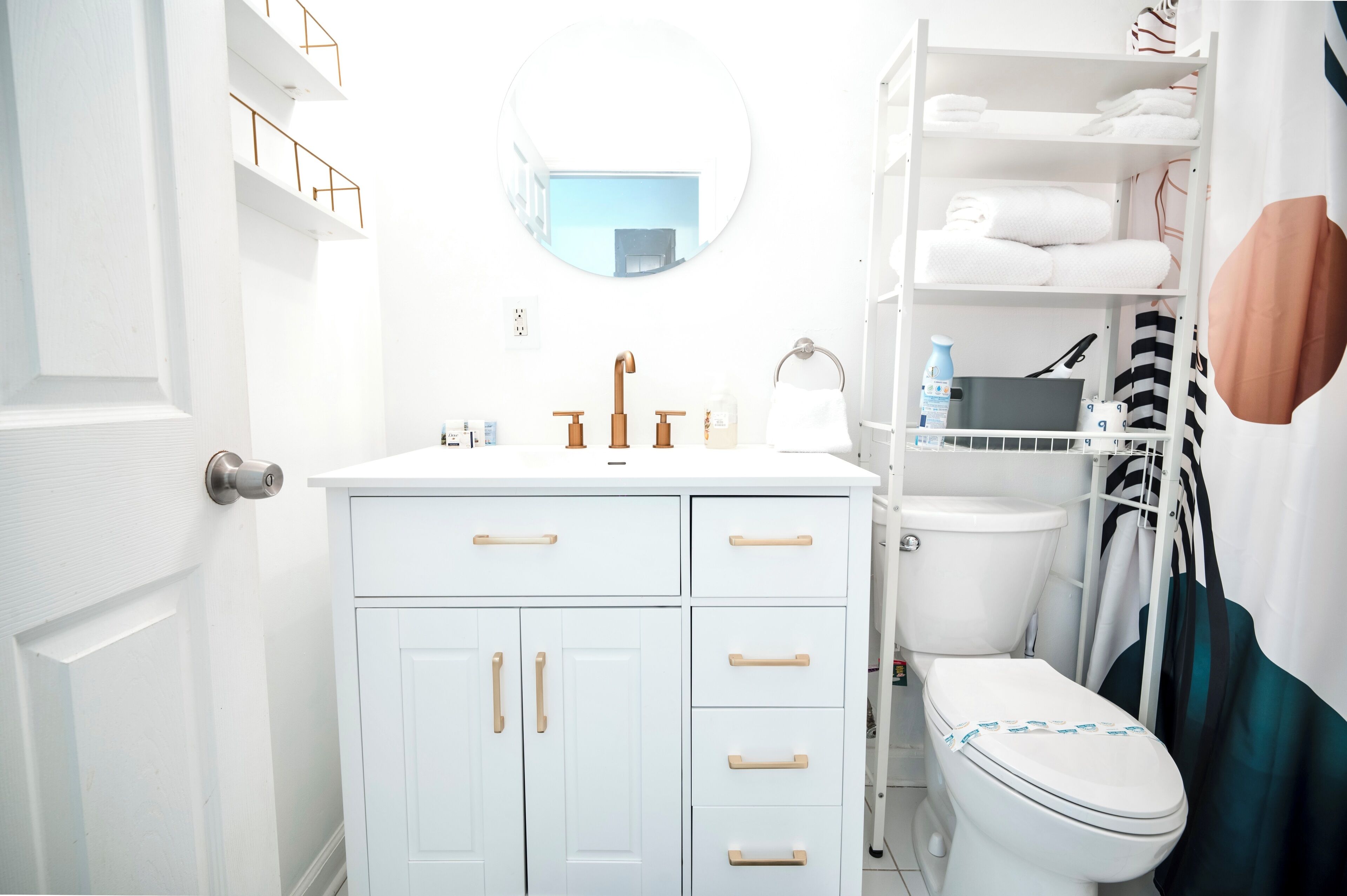 Et hvidt badeværelse med et rundt spejl, en hvid vask og et toilet. Der er hylder med håndklæder og toiletartikler.