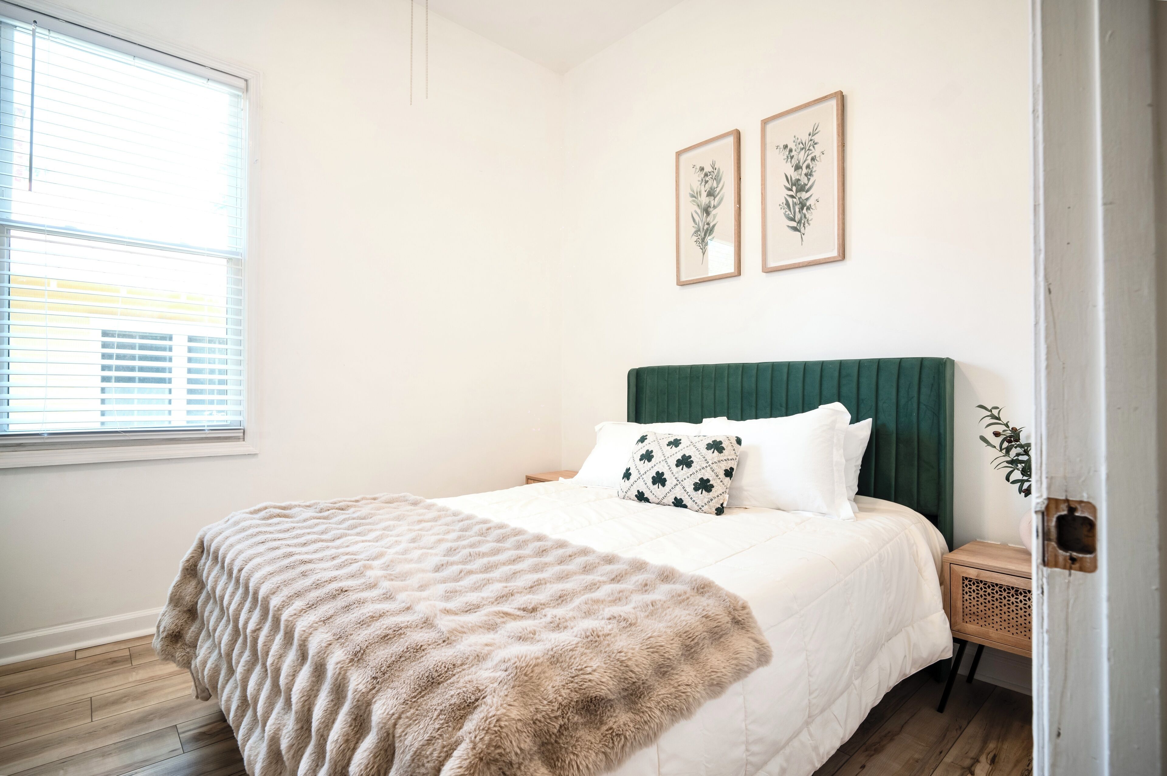 A bedroom with a bed, a green headboard, a wooden nightstand, and two framed botanical prints on the wall.