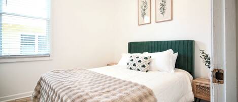 A bedroom with a bed, a green headboard, a wooden nightstand, and two framed botanical prints on the wall.