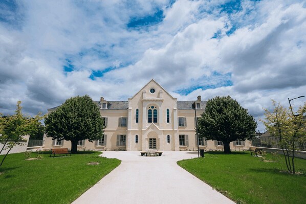 A large, symmetrical building with a central entrance, flanked by two large trees and a bench in the foreground.