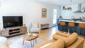 A modern living room with a large TV, comfortable seating, a small coffee table, and a kitchen area with bar stools. - 5 Min to Downtown Warwick, Wineries, Breweries, Hikes, Shops, & Greenwood Lake! (Warwick)