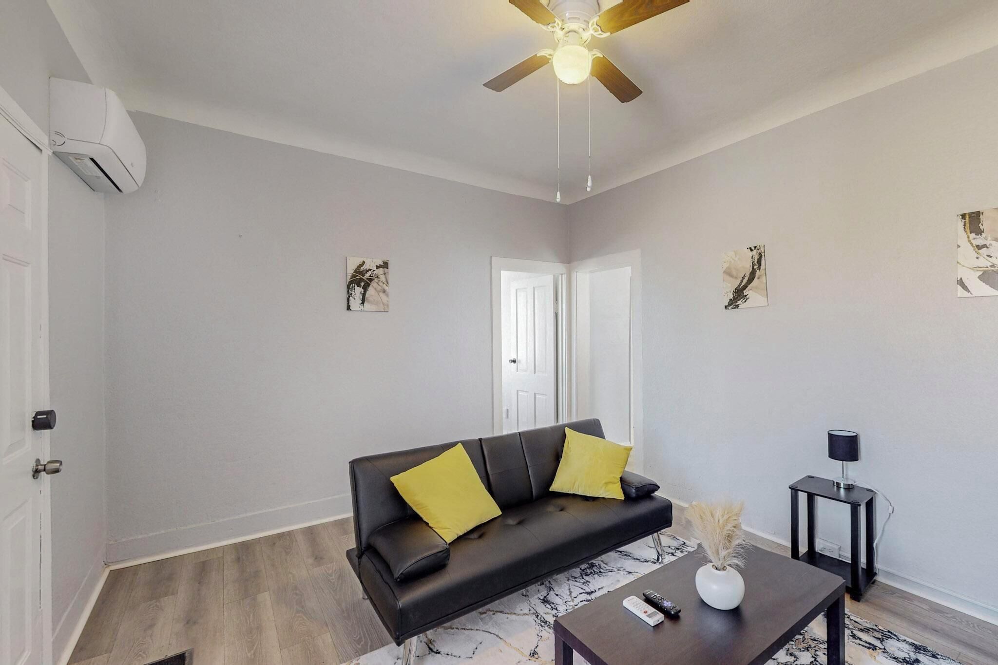 Un salon avec un canapé en cuir noir, deux coussins jaunes, un ventilateur de plafond et une petite table sur laquelle est posé un vase.