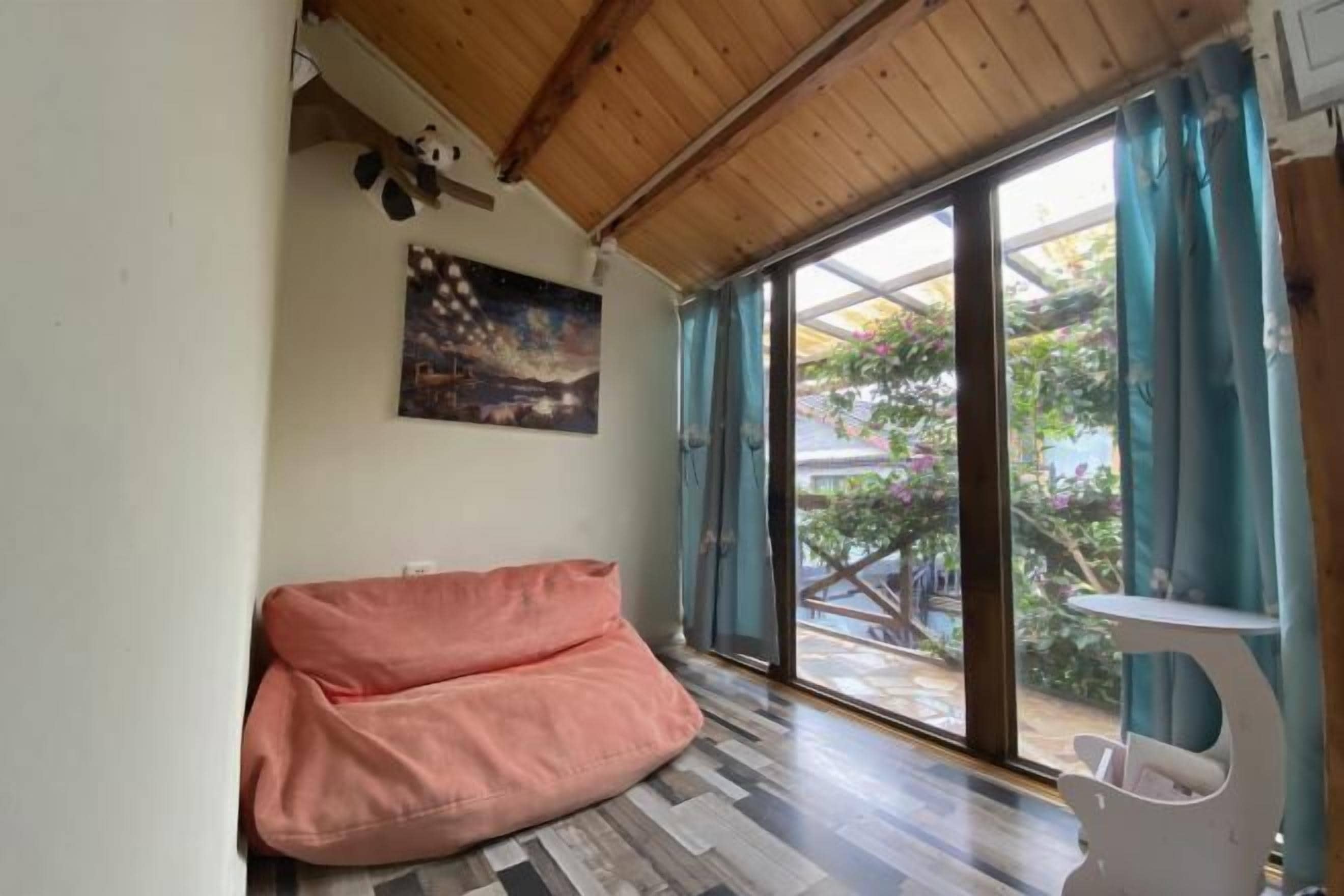 A room with a wooden ceiling, a pink bean bag chair, a white table, and a large window with curtains.