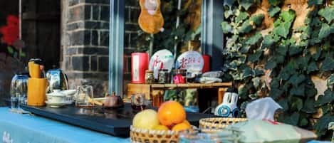 Una mesa dispuesta con diversos objetos, incluyendo una cesta, una botella y algunos platos, frente a paredes cubiertas de hiedra y una estructura de ladrillo.