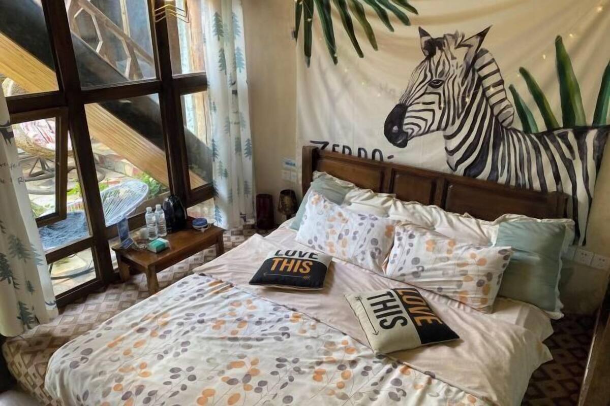 Una cama con un tapiz de cebra, almohadas con el texto 'LOVE THIS' y una ventana que da a una hamaca y follaje.