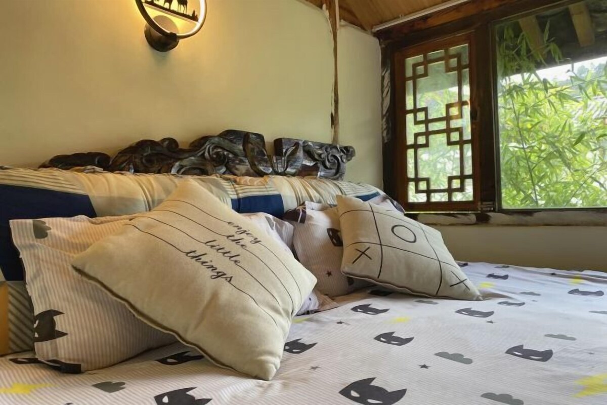 A bed with decorative pillows, a wall-mounted light, and a window with a view of greenery.