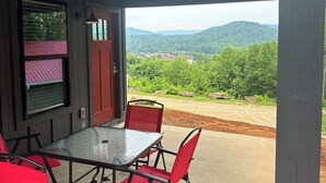 A covered patio with a table and chairs, a red door, and a view of a forested area. - Nacoochee 3, Cozy cabin, close to town, walking trails, winery, adventures  (Helen)