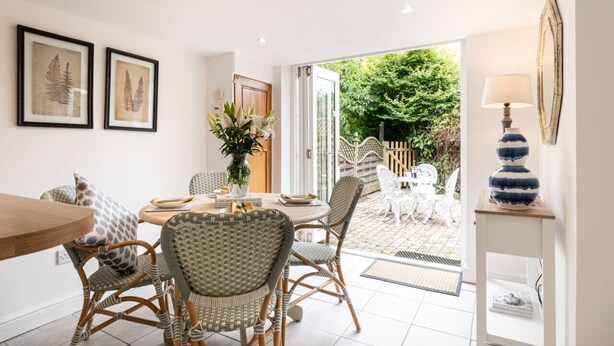 A dining area with wicker chairs, a wooden table, and a vase of flowers. There are framed pictures on the wall and a lamp on a side table. - Poppy Cottage - Cosy 2BD Home, Perfect For Family (Clifford Chambers)