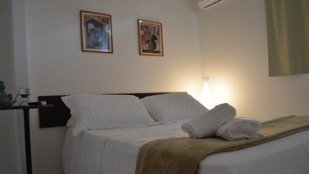A neatly made bed with white linens and a brown throw blanket, a bedside table with a lamp, and two framed pictures on the wall. - POUSADA DAS COLÔNIAS ILHA DA GIJÓIA (Rio de Janeiro)