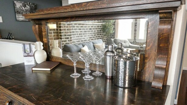 Una barra de madera oscura con un espejo encima que refleja una pared de ladrillo y una ventana. Sobre la barra, varios vasos de vino transparentes, una jarra de metal y un libro pequeño.