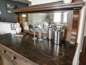 A dark wooden bar counter with a mirror above it, reflecting a brick wall and a window. On the counter, there are several clear wine glasses, a metal pitcher, and a small book.