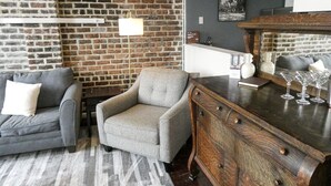 Una sala de estar con pared de ladrillo, un sofá gris, un sillón gris, un mueble de madera y una zona de bar con vasos.
