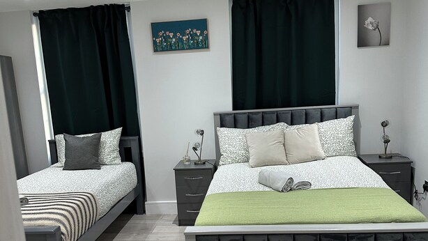 A modern bedroom with two single beds, a large bed, a bedside table with a lamp, and a green throw on the large bed. - Guest&Rest Elite  (London)