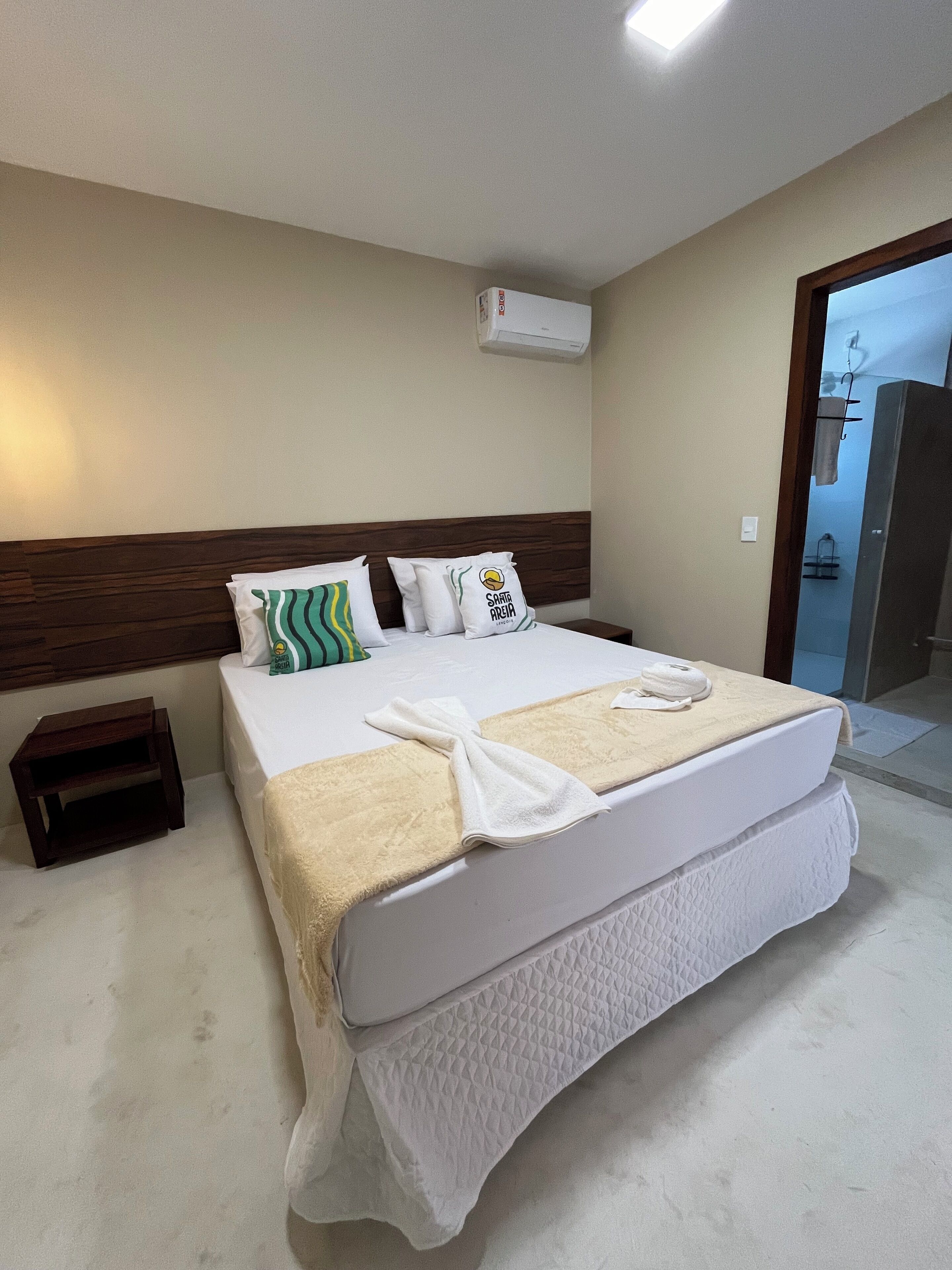 Una habitación de hotel con una cama grande, cabecera de madera, mesita de noche pequeña y un equipo de aire acondicionado en la pared.
