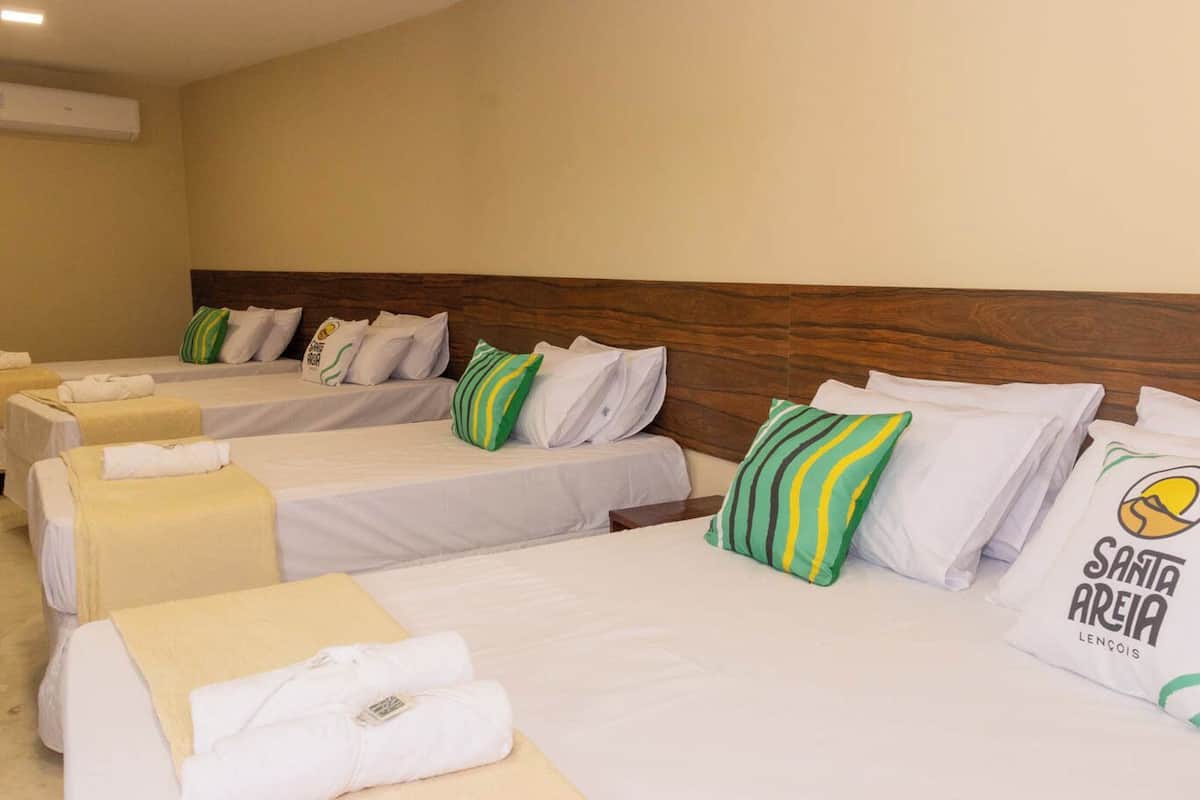 A hotel room with four beds, each with white bedding and green striped pillows. There is a wooden headboard, and a wall-mounted air conditioner is visible.