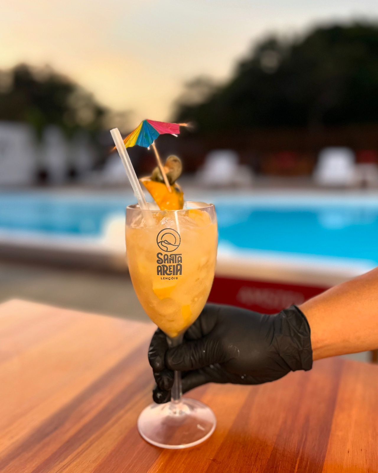 Un verre à cocktail, un petit parasol tropical et une tranche de fruit sont posés sur une table en bois, près d’une piscine.