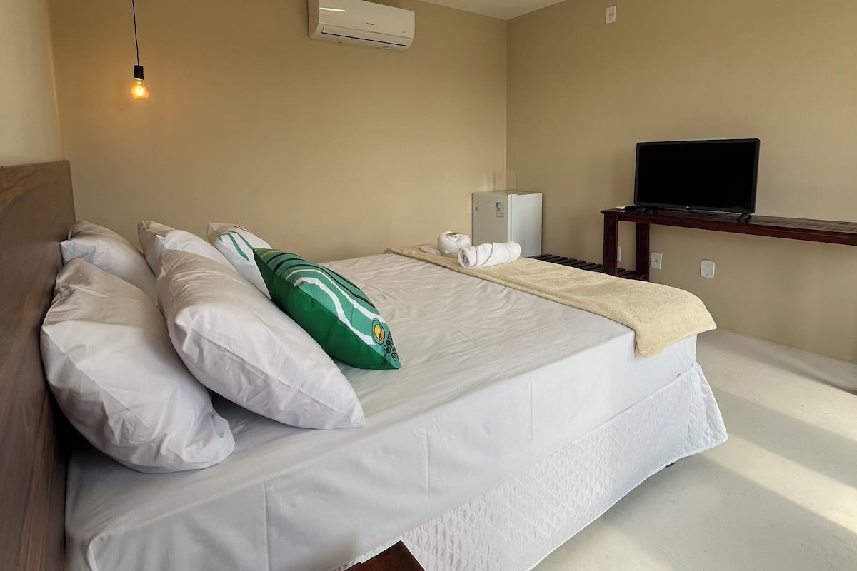 A hotel room with a bed, pillows, a bedside table, a TV, an air conditioner, and a ceiling light.