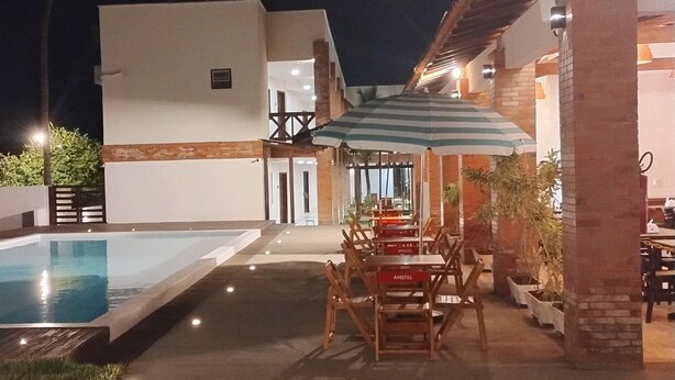 An outdoor pool area with a dining setup under a striped umbrella, surrounded by potted plants and a building with a modern design. - Santa Areia Lençóis Pousada (Santo Amaro do Maranhão)
