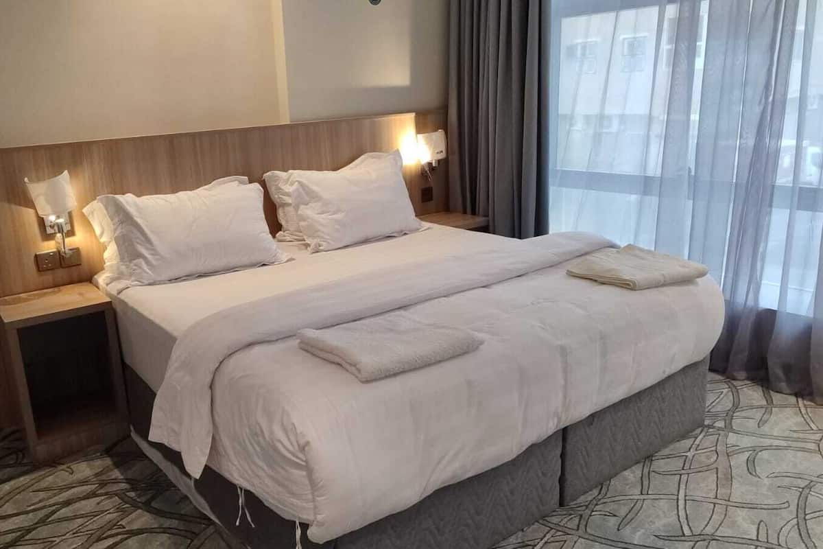 A hotel room with a large bed, two bedside tables with lamps, and a view of the city through sheer curtains.