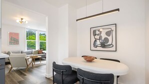 Ein moderner Essbereich mit einem weißen Tisch und dunklen Stühlen, über dem ein gerahmtes abstraktes Kunstwerk hängt, und einem Fenster mit Blick auf Grünflächen.