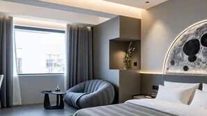 Una habitación de hotel moderna con una cama grande, una zona de estar con una silla, una mesita pequeña y una obra de arte colgada en la pared.