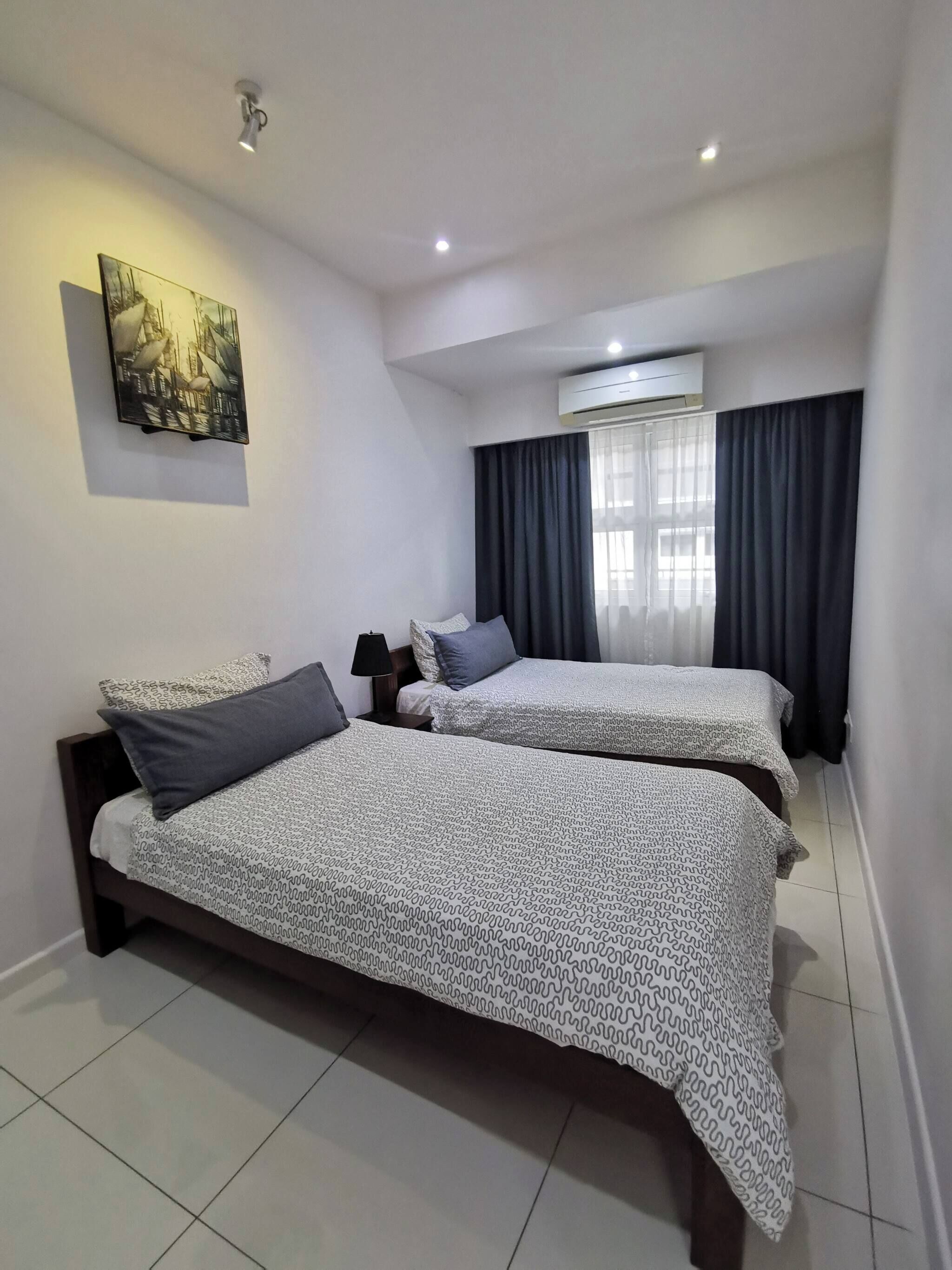 A hotel room with two beds, a ceiling light, an air conditioning unit, and a painting on the wall.
