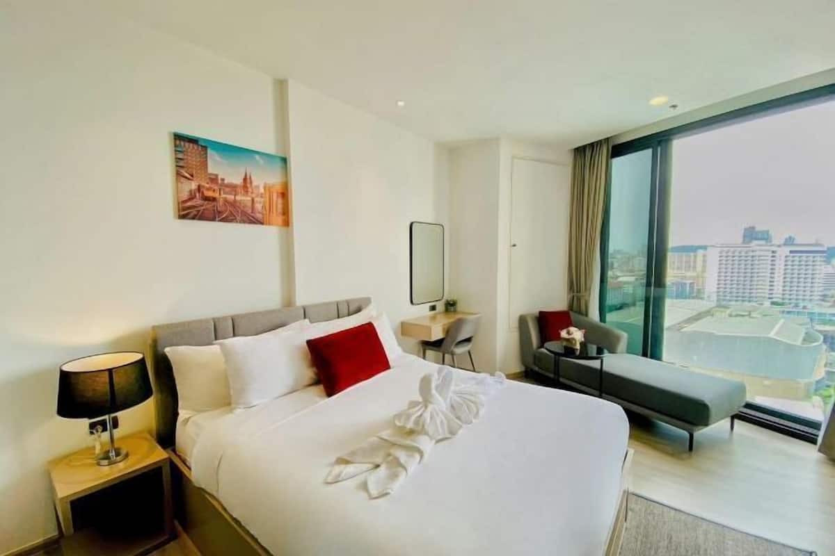 A hotel room with a bed, a desk, a chair, a large window with a city view, and a painting on the wall.