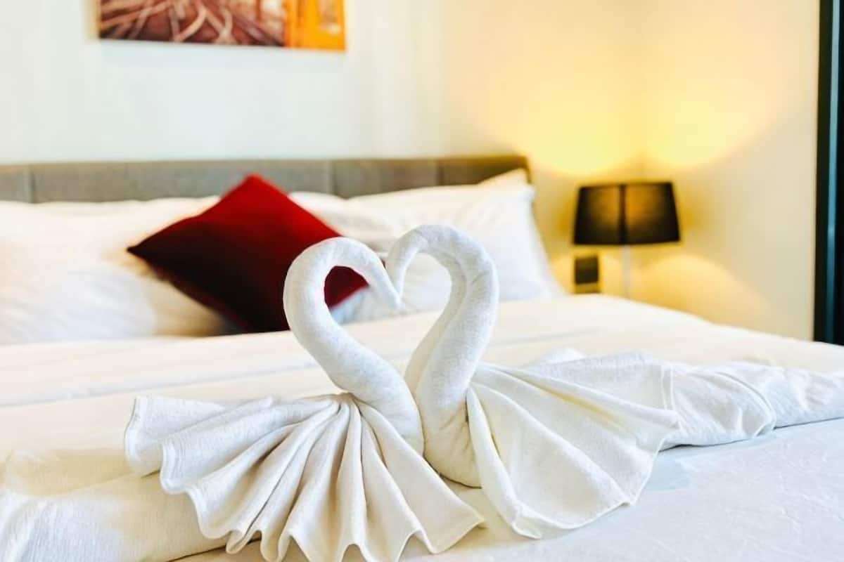 A bed with a towel arranged in the shape of two swans, a red pillow, and a lamp on the nightstand.
